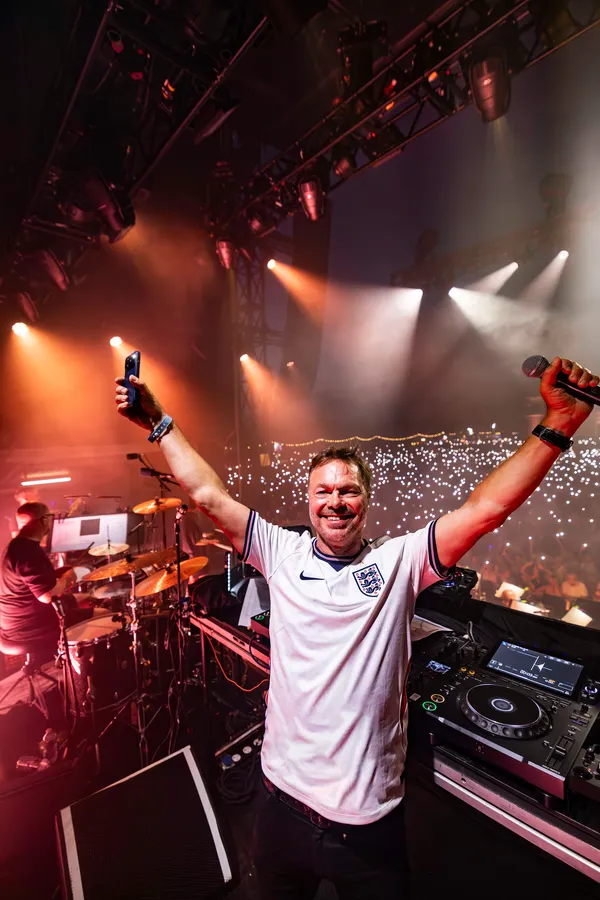 Pete Tong in an England football shirt, arms raised on stage under red and white lights